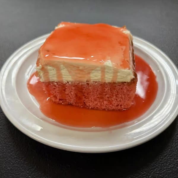 A slice of pink cake with white frosting and a red glaze sits on a white plate.